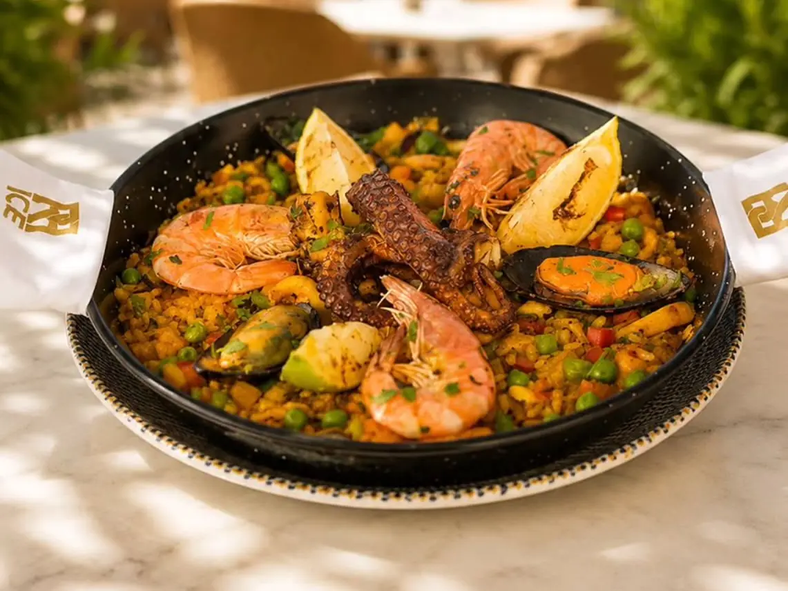 Notre paëlla aux fruits de mer servie sur une table en marbre de la terrasse, poulpe, crevettes et moules, les serviettes au monogramme doré nouées aux anses