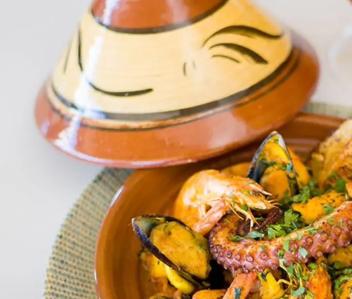 La coiffe en terre peinte du tajine de la mer, posée à côté du plat, crevettes et poulpe au premier plan